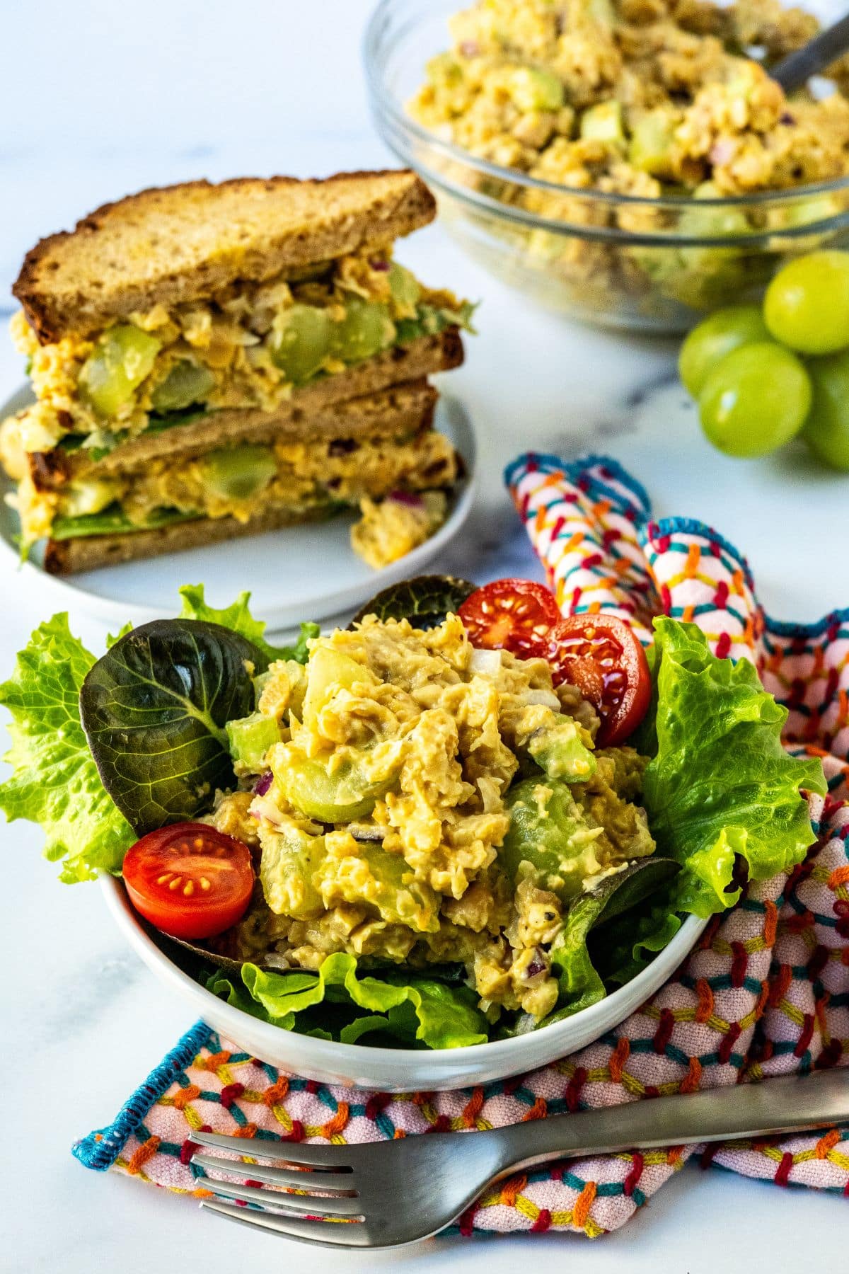 A bowl of salad topped with tomatoes and chickpea salad, a chickpea salad sandwich, and a mixing bowl with curried chickpea salad in it.