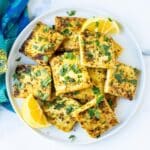Crispy squares of lemon pepper tofu garnished with fresh parsley on a platter with lemon wedges,