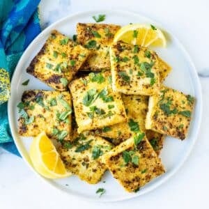 Crispy squares of lemon pepper tofu garnished with fresh parsley on a platter with lemon wedges,