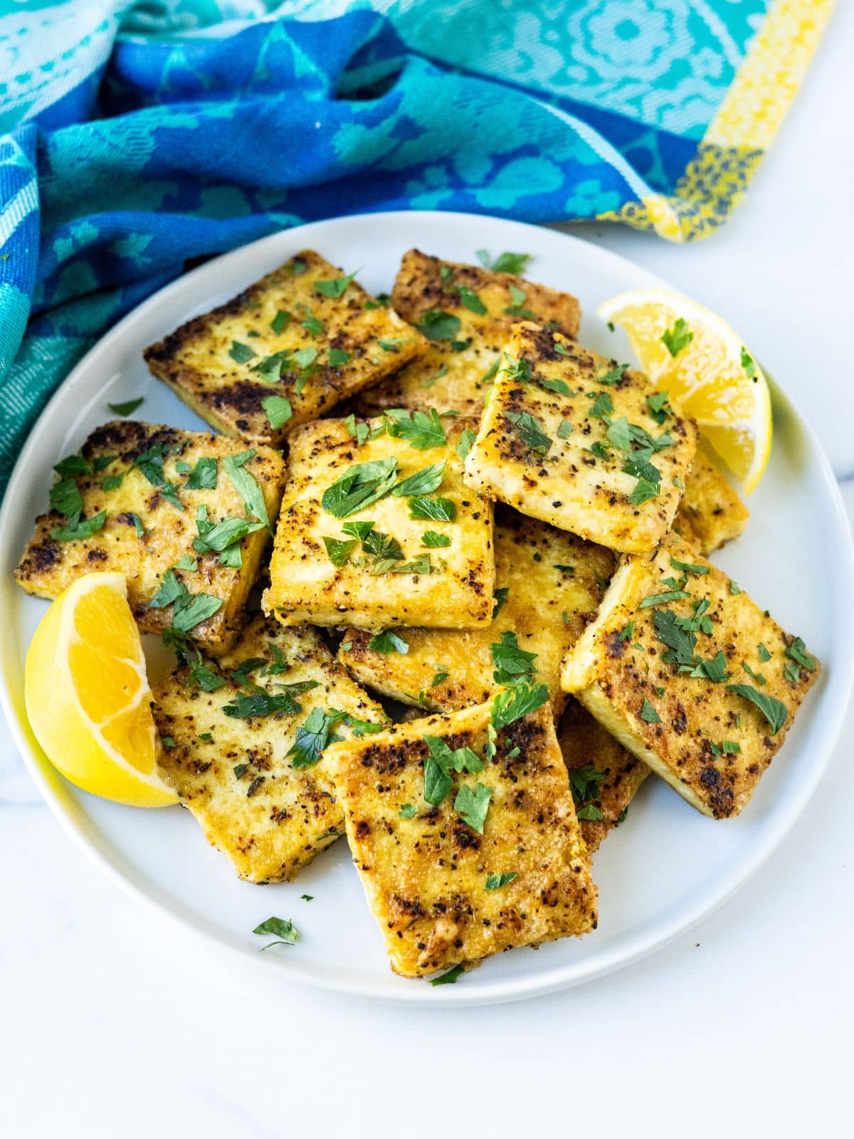 Squares of crispy, browned tofu garnished with fresh parsley on a plate with lemon wedges.