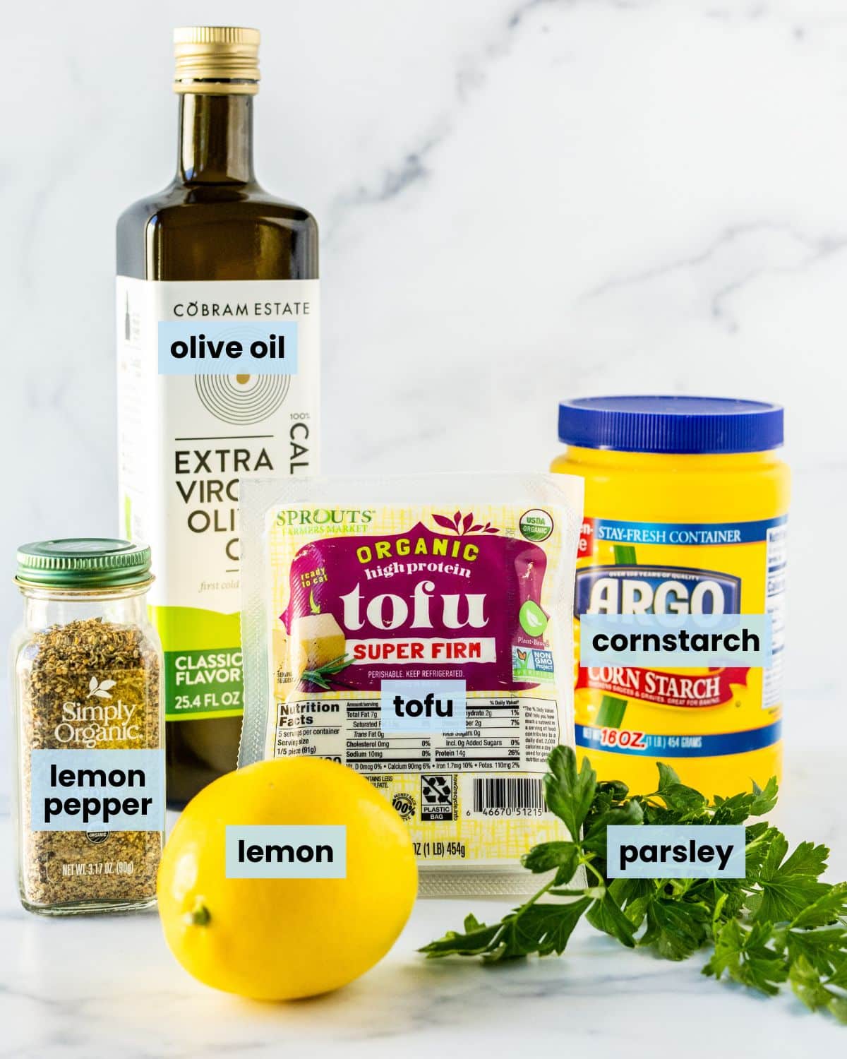 A lemon, a small bunch of fresh parsley, a jar of lemon pepper seasoning, a bottle of olive oil, a block of tofu, and a container of cornstarch.