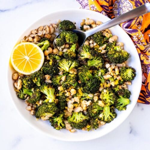 Bowl of roasted broccoli and white beans garnished with lemon zest and a slice of lemon on the side.