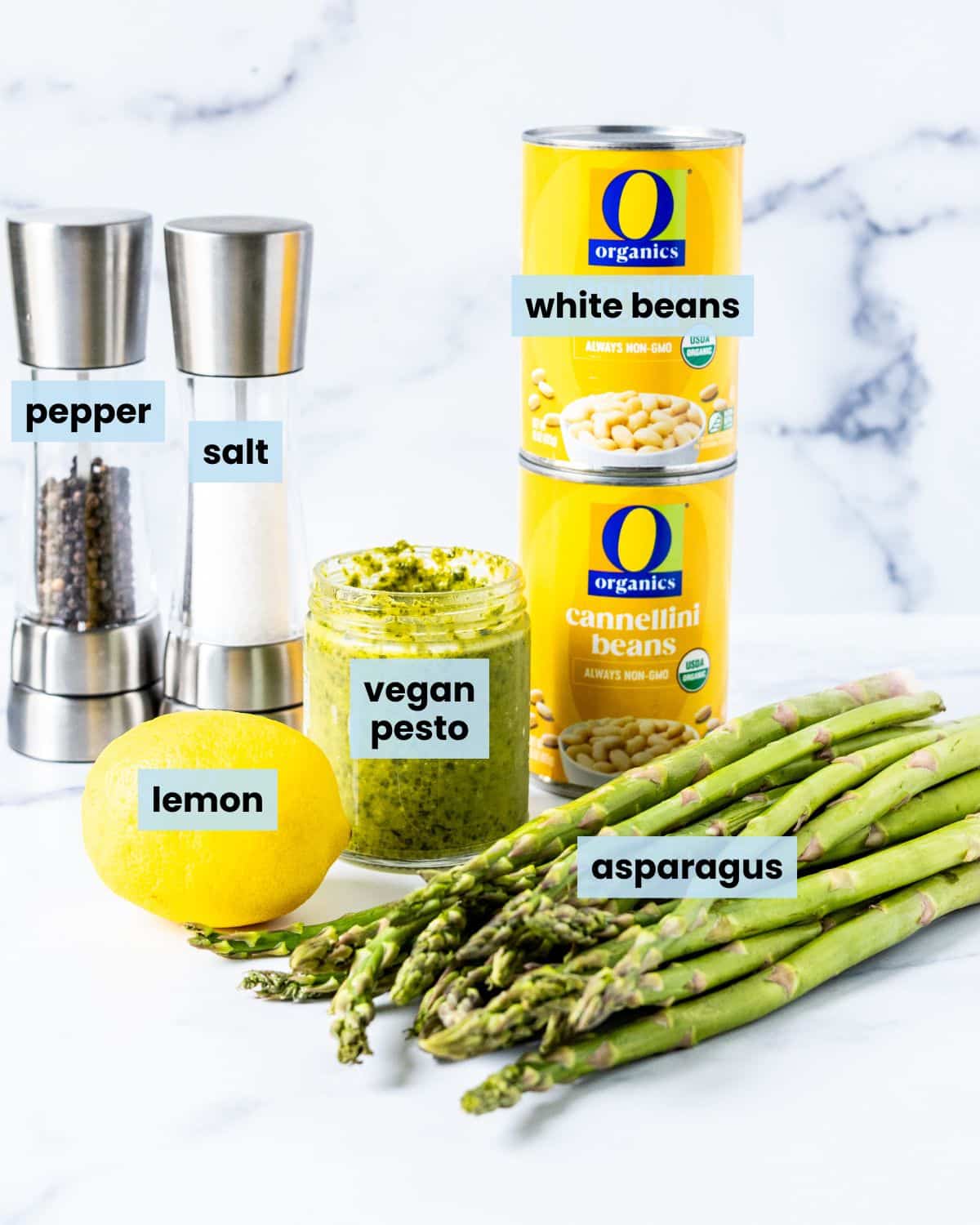 Bunch of asparagus, a lemon, a jar of vegan pesto, two cans of white beans, and salt and pepper grinders on a countertop.