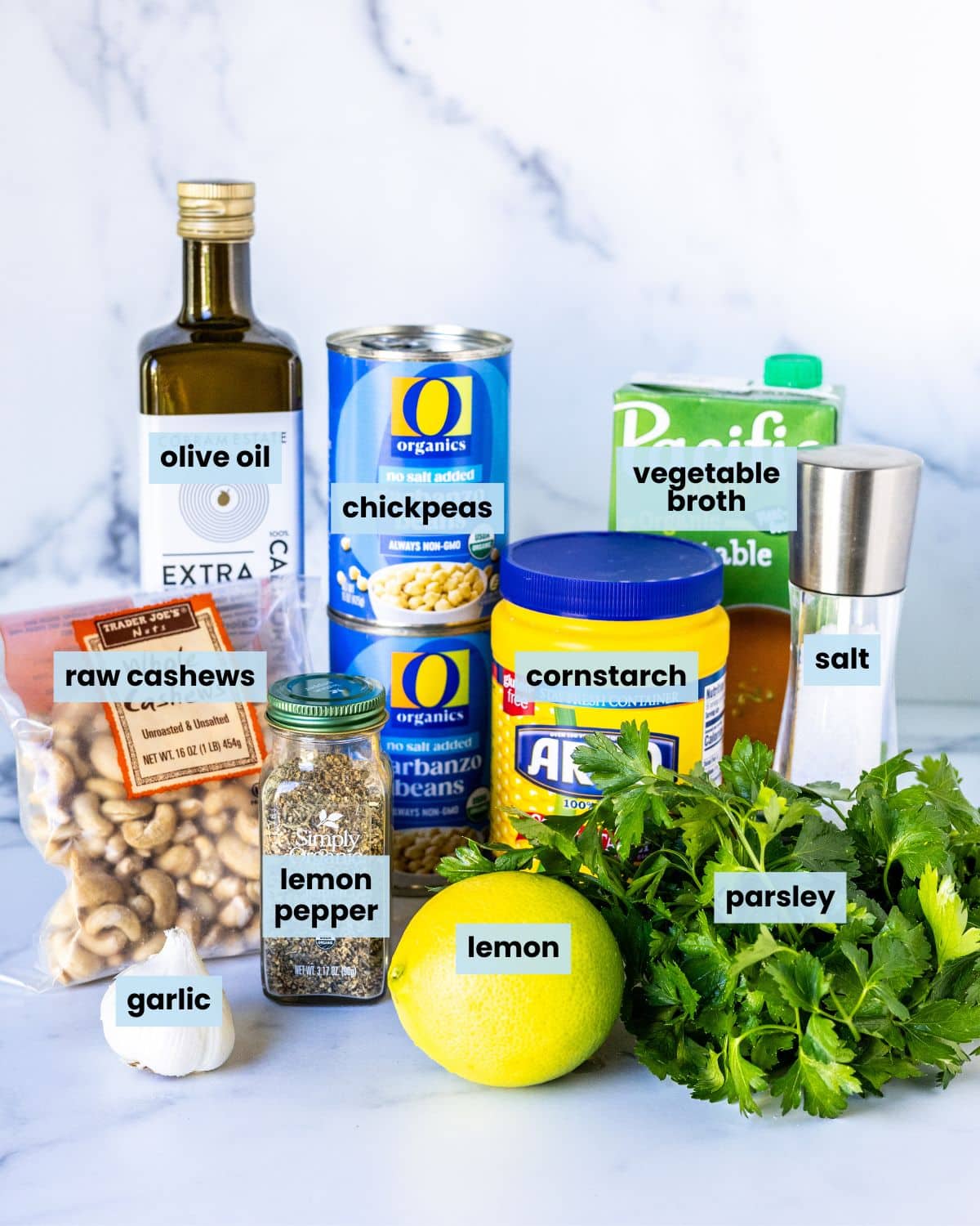 Fresh garlic, lemon, and parsley; bag of raw cashews; jar of lemon pepper seasoning; container of cornstarch; salt grinder; bottle of olive oil; two cans of chickpeas; and a carton of vegetable broth on a kitchen counter.