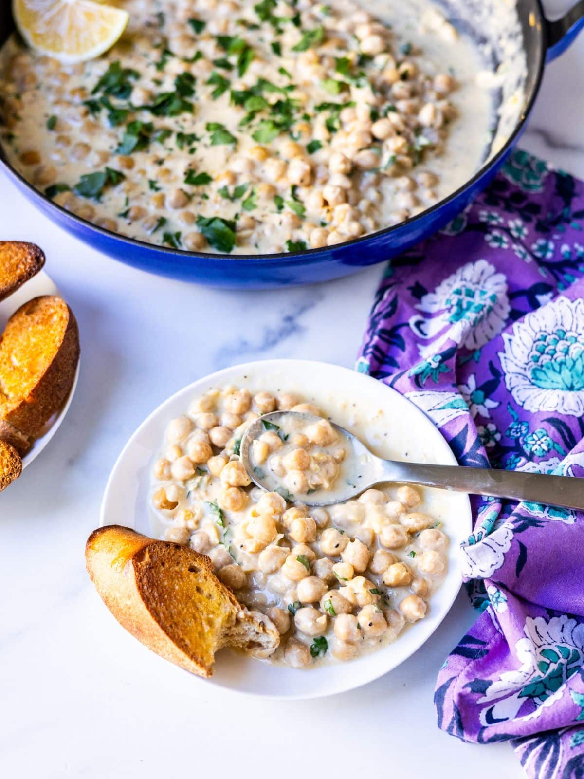 A plate with a spoon in a serving of lemon pepper chickpeas and a bite taken out of a slice of baguette, with the skillet in the background.
