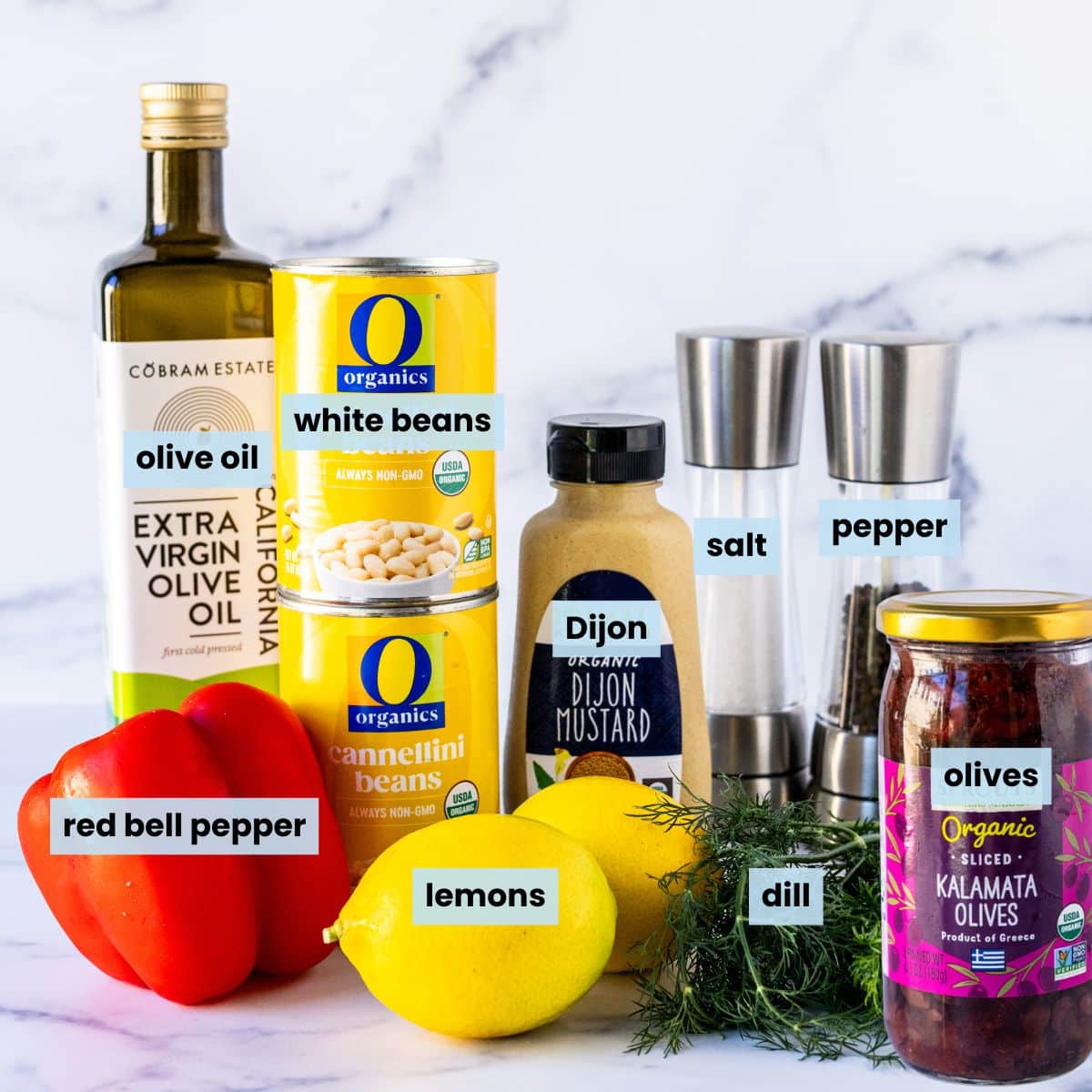 A red bell pepper, two lemons, a small bunch of fresh dill, a jar of Kalamata olives, a bottle of olive oil, two cans of white beans, a bottle of Dijon mustard, and salt and pepper grinders.