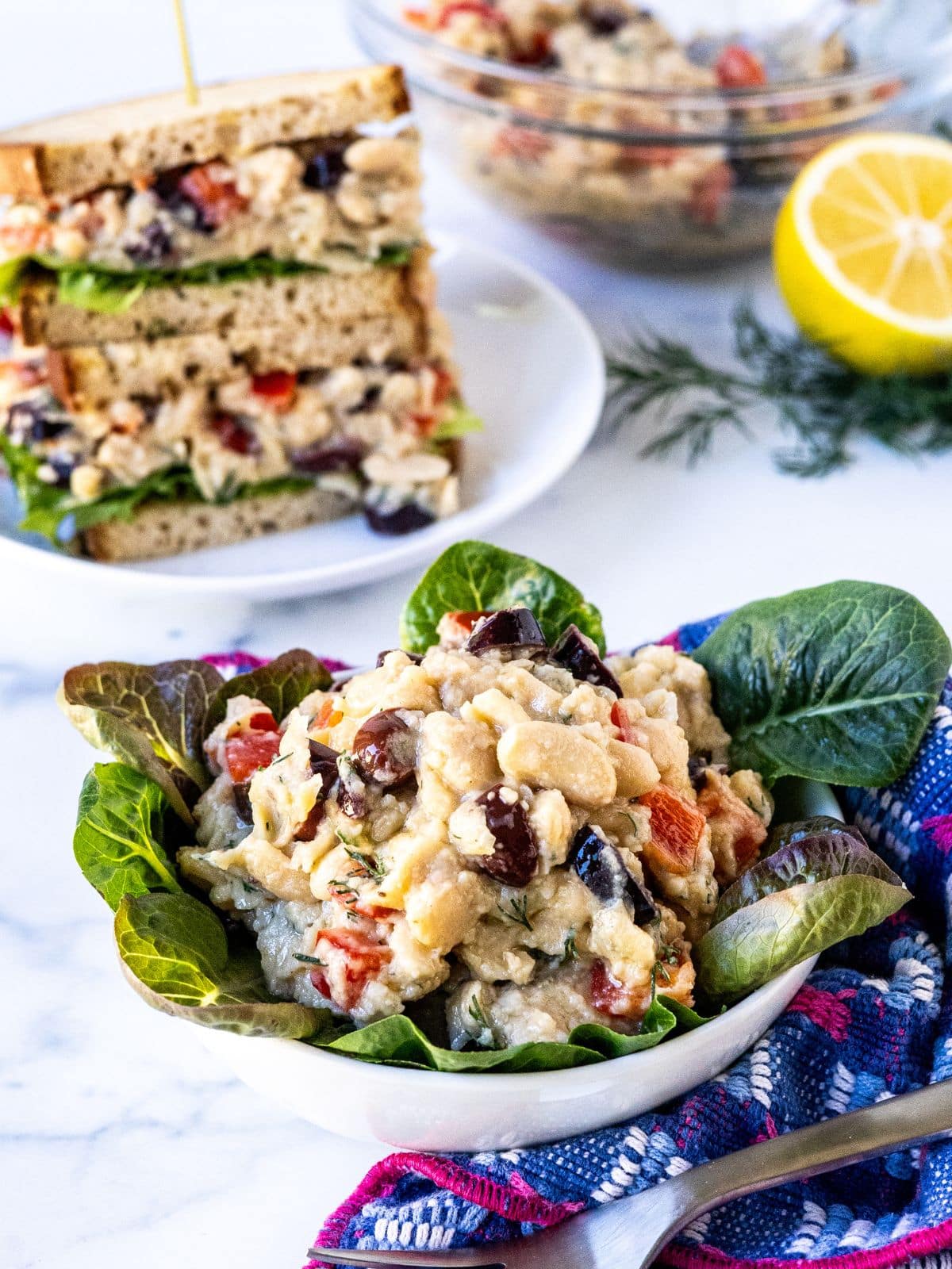 Scoops of smashed white bean salad on a bed of lettuce, a sandwich cut in half, and a mixing bowl with the white bean salad next to fresh dill fronds and half a lemon.