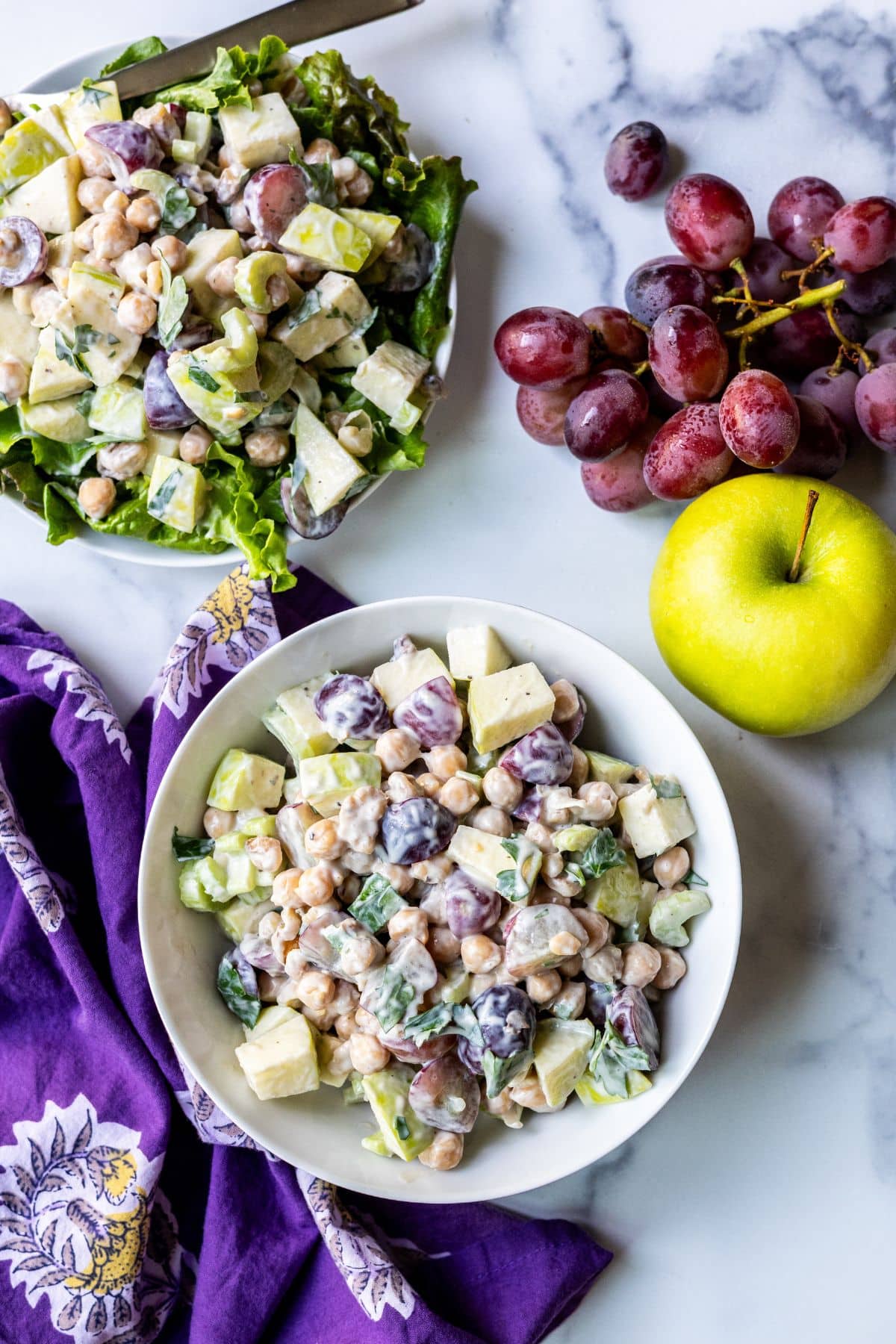 Bowl of chickpea Waldofr salad, a serving of the salad on a bed of lettuce, a green apple, and a bunch of red grapes.