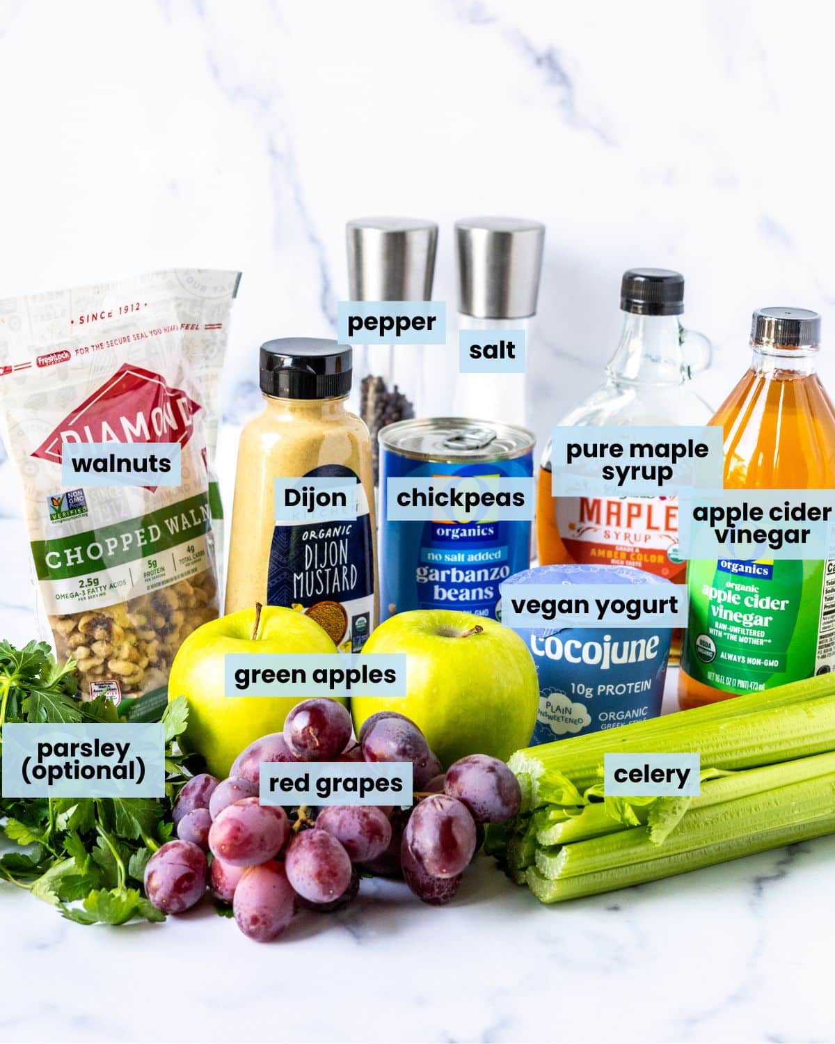 Ingredients on a kitchen counter: fresh parsley, red grapes, a bunch of celery, two green apples, a bag of chopped walnuts, a jar of Dijon mustard, a can of chickpeas, a container of plain vegan yogurt, a bottle of apple cider vinegar, a bottle or pure maple syrup, and salt and pepper grinders.