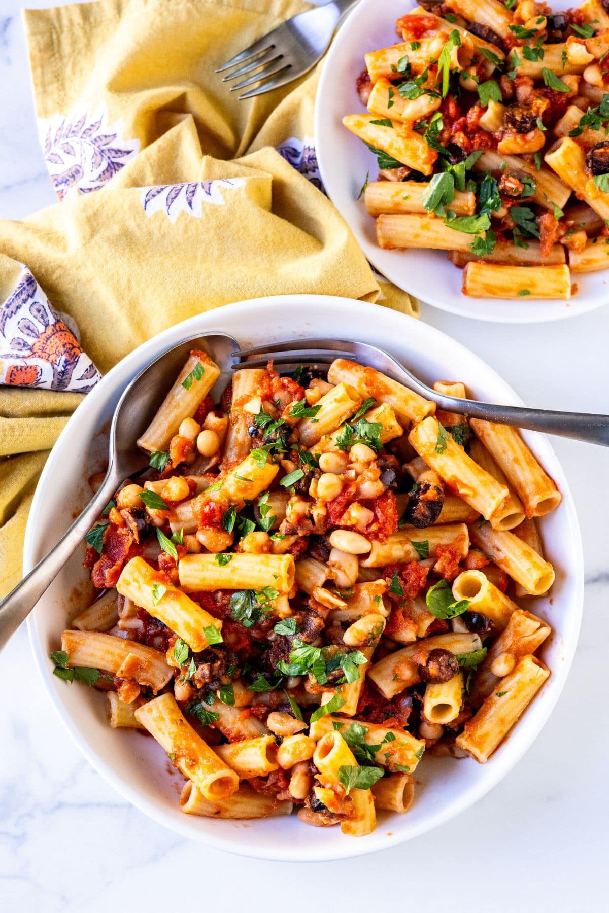 Bowl of white bean tomato pasta with olives and a individual serving on a plate.