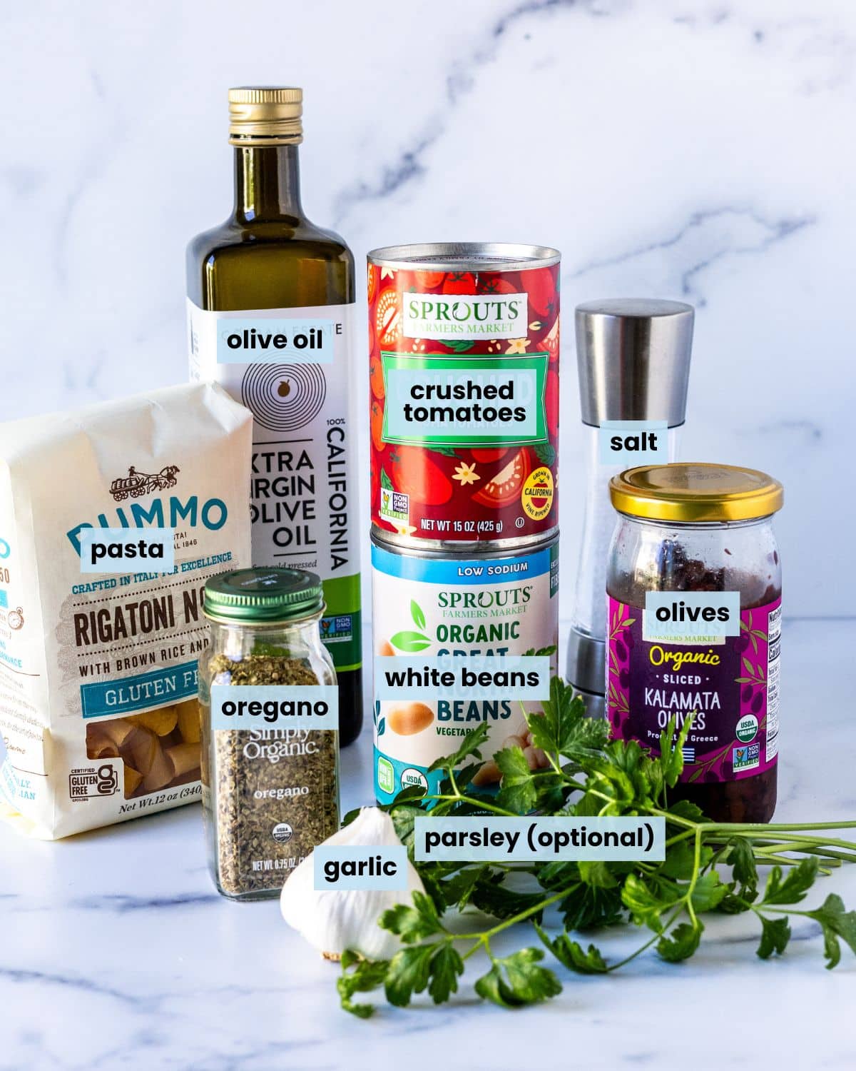 Head of garlic, fresh parsley, bag of rigatoni, jar of dried oregano, bottle of olive oil, cans of crushed tomatoes and white beans, salt, and a jar of Kalamata olives on a kitchen counter.
