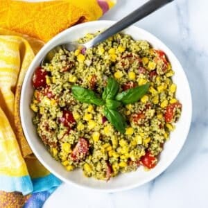Bowl of vegan pesto quinoa salad with corn and tomatoes garnished with a sprig of fresh basil.