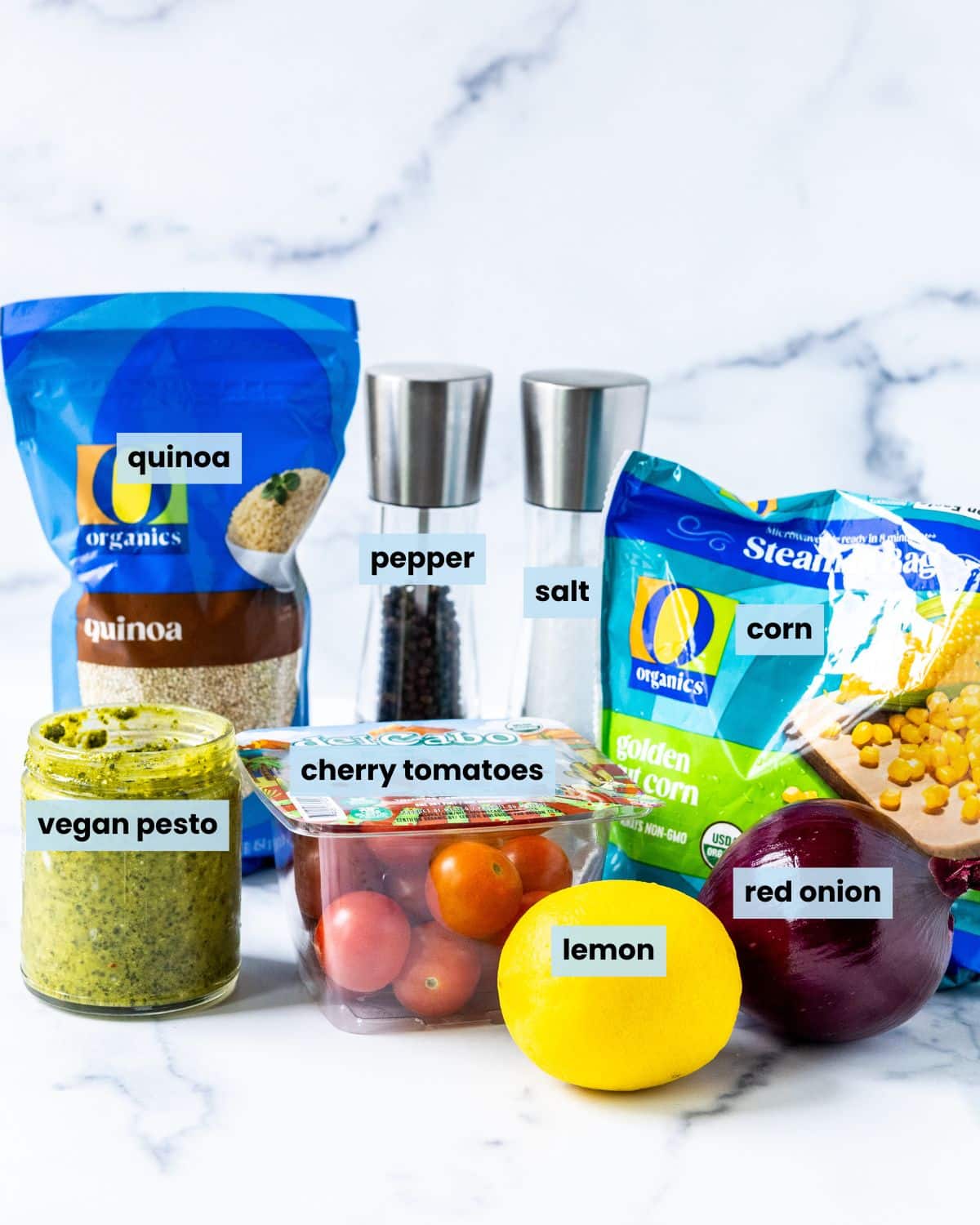 Bag of quinoa, jar of vegan pesto, container of cherry tomatoes, a lemon, a red onion, a bag of frozen corn, and salt and pepper grinders on a kitchen counter.