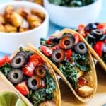 Potato and kale tacos topped with tomatoes, black olives, and hot sauce with a slice of lime on the side.