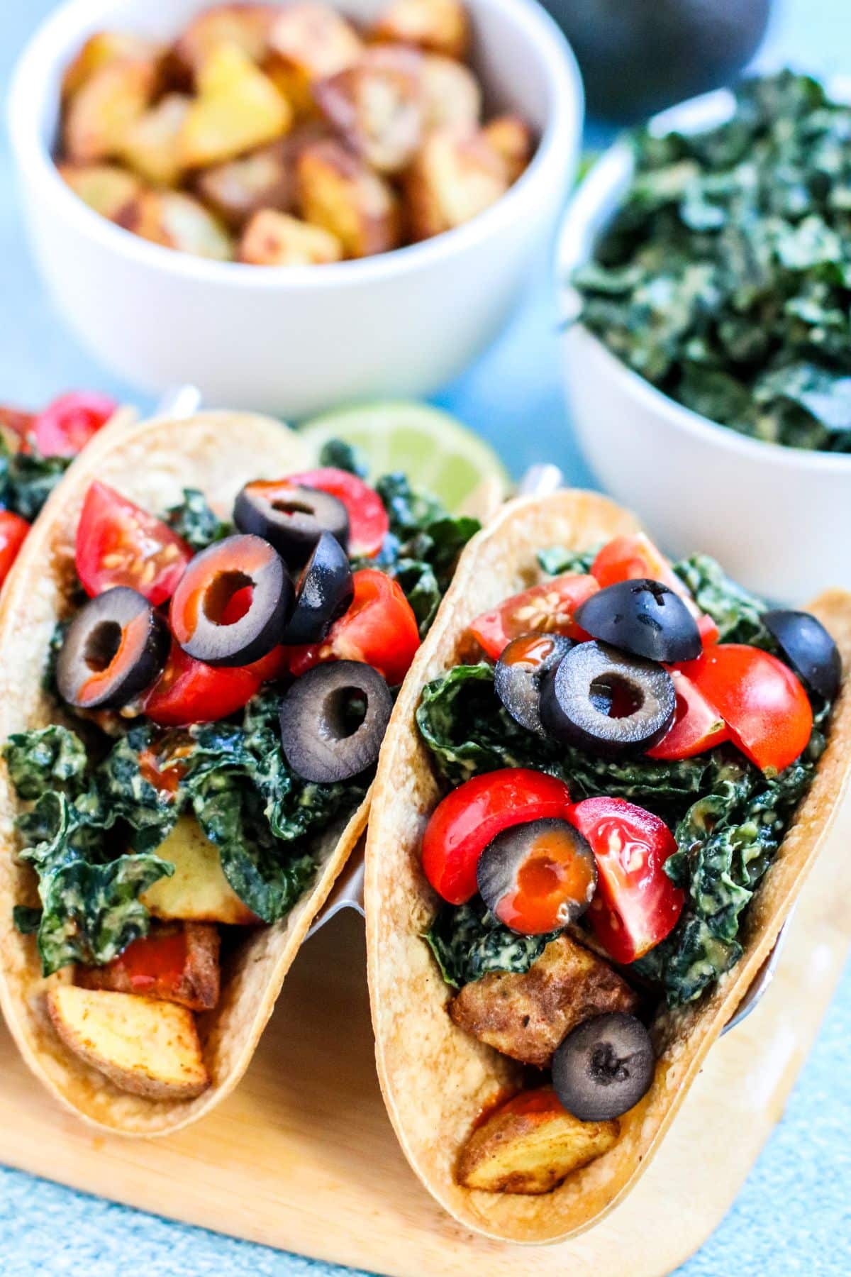 Two tacos in front of bowls of roasted potatoes and massaged kale.