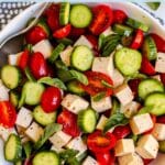 Bowl of salad made with sliced cucumbers, halved cherry tomatoes, fresh basil leaves, and tofu feta cubes, with text overlay: Tomato Basil Cucumber Salad with Vegan Feta.