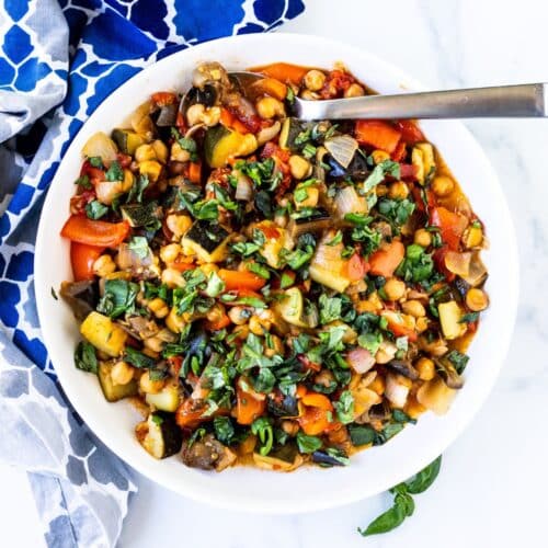Spoon in a bowl of roasted chickpea ratatouille garnished with fresh basil.