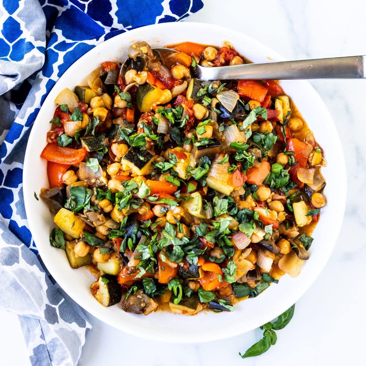 Spoon in a bowl of roasted chickpea ratatouille garnished with fresh basil.