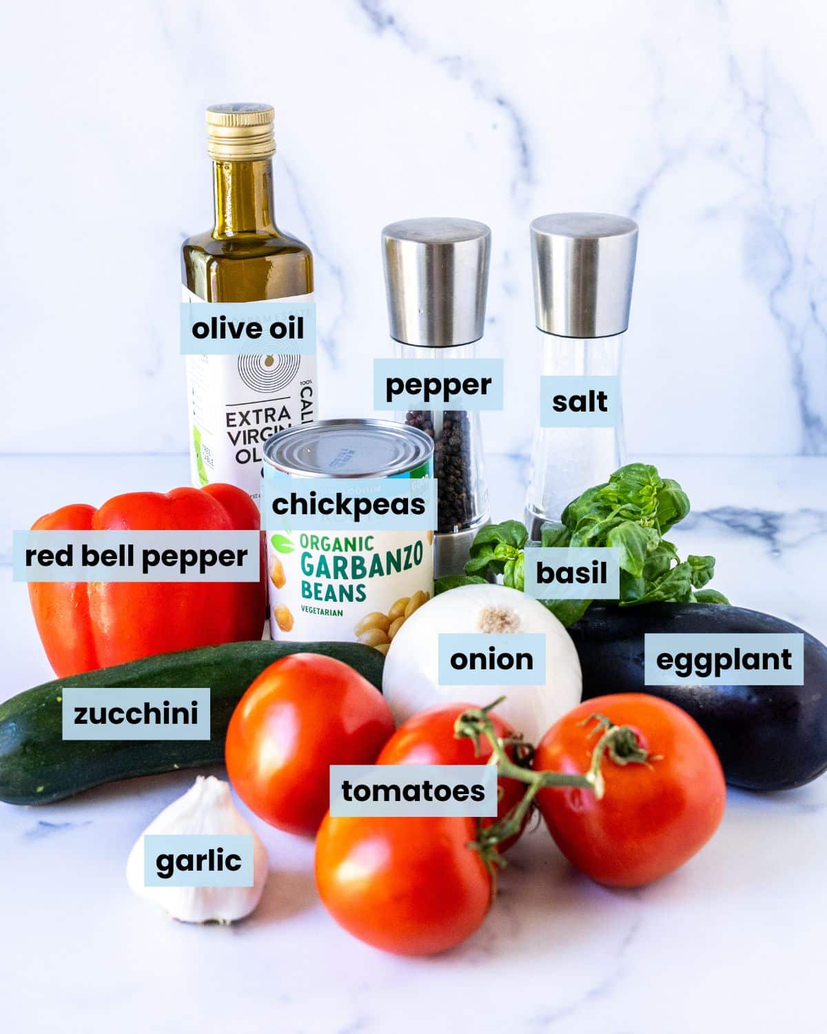 Ingredients on a kitchen counter including zucchini, eggplant, tomatoes, red bell pepper, chickpeas, onion, garlic, basil, olive oil, salt, and pepper.