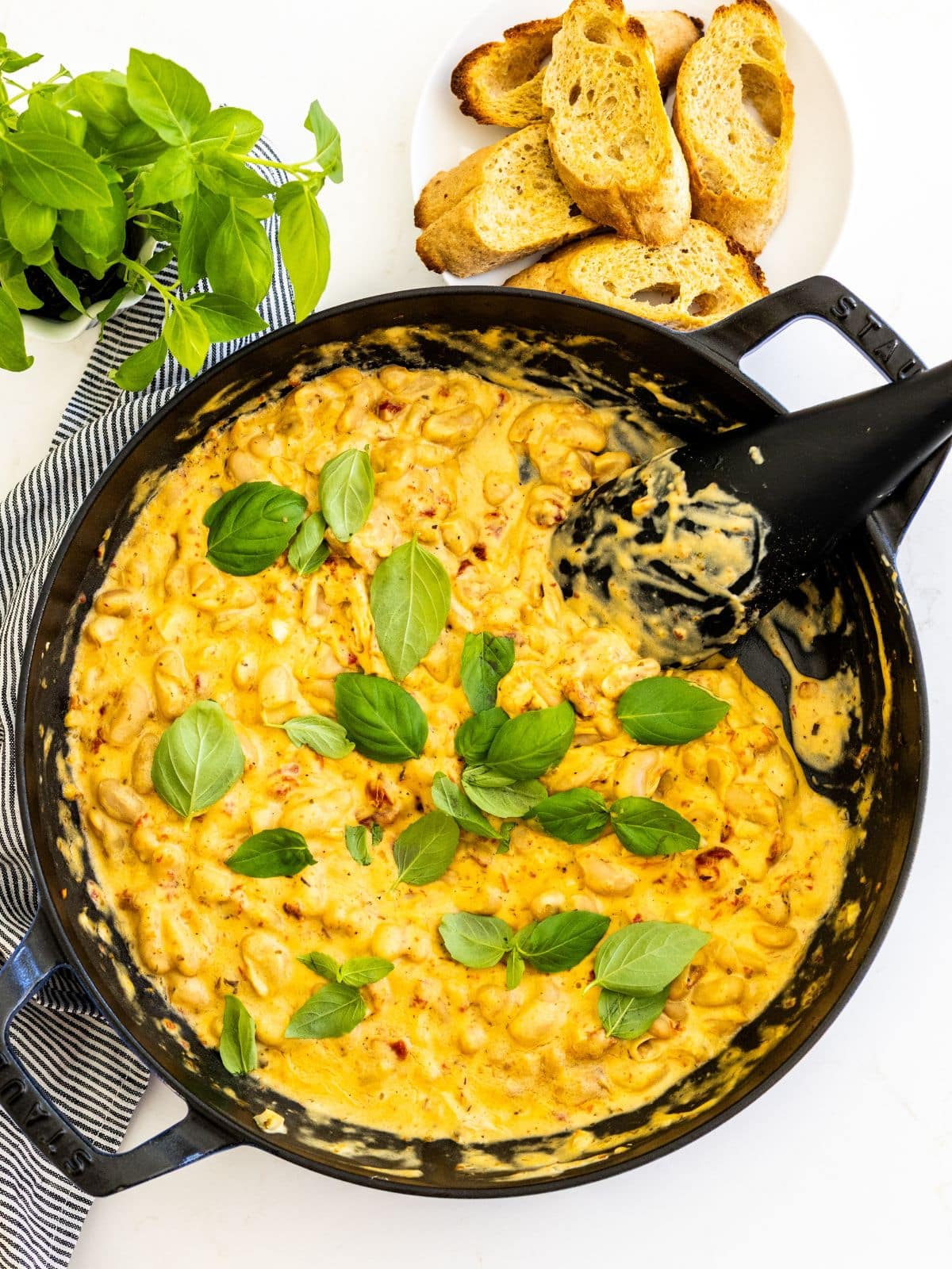 Spoon in a skillet of creamy white beans with sun-dried tomatoes garnished with fresh basil leaves alongside a plate of toasted baguette slices.