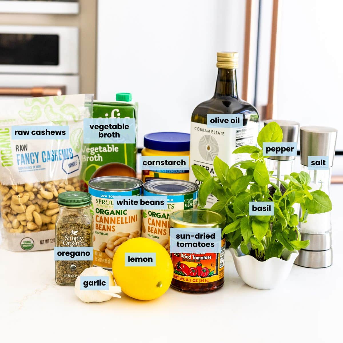 Recipe ingredients on a kitchen counter: garlic, lemon, jar of dried oregano, jar of sun-dried tomatoes in olive oil, cans of white beans, fresh basil, bag of raw cashews, carton of vegetable broth, canister of cornstarch, bottle of olive oil, and salt and pepper grinders.