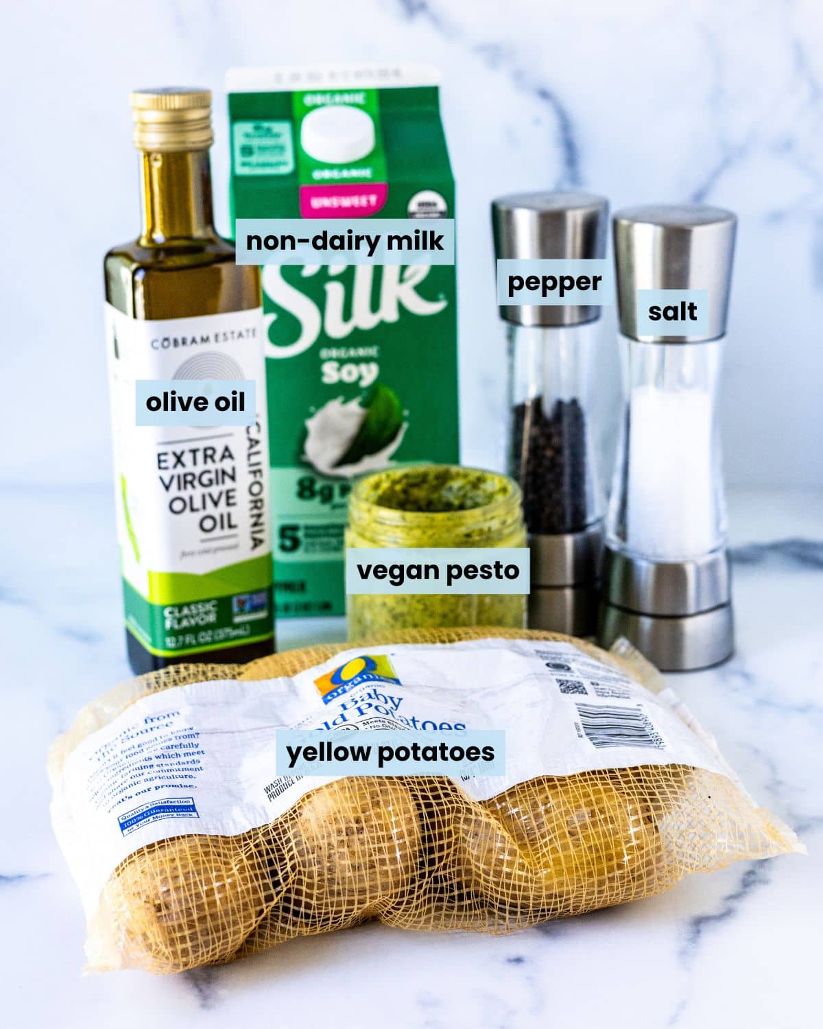 Bag of yellow potatoes, jar of olive oil, carton of non-dairy milk, jar of pesto, and salt and pepper grinders on a kitchen counter.