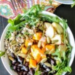 Bowl with arugula, quinoa, roasted butternut squash, black beans, sliced apples,, and pumpkin seeds, drizzled with tahini dressing, with text overlay Butternut Squash Quinoa Bowls.